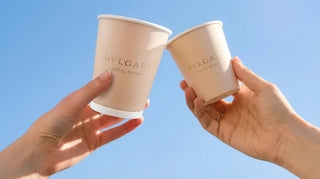 Two BVLGARI Hotel Roma branded paper cups held up against a clear blue sky